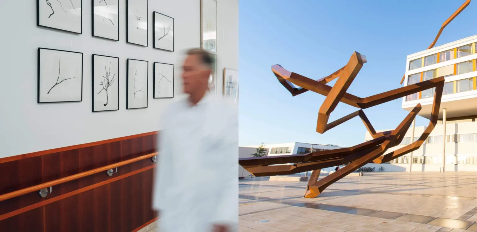 Arzt geht an einer Bildwand eines Klinikflures vorbei und Metallkunstwerk vor einem Klinikgebäude