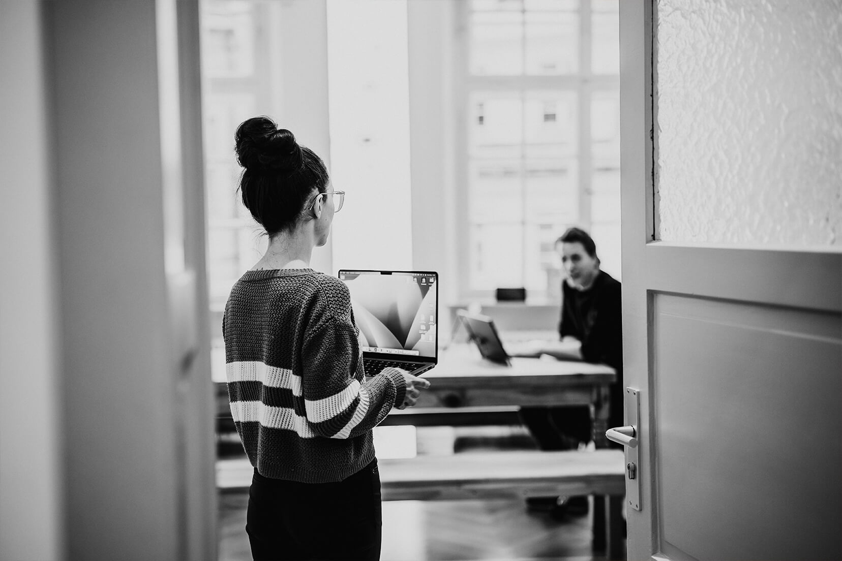 Eine Mitarbeiterin mit einem laptop in der Hand steht im Türahmen zu einem Raum in dem ein weiterer Mitarbeiter sitzt