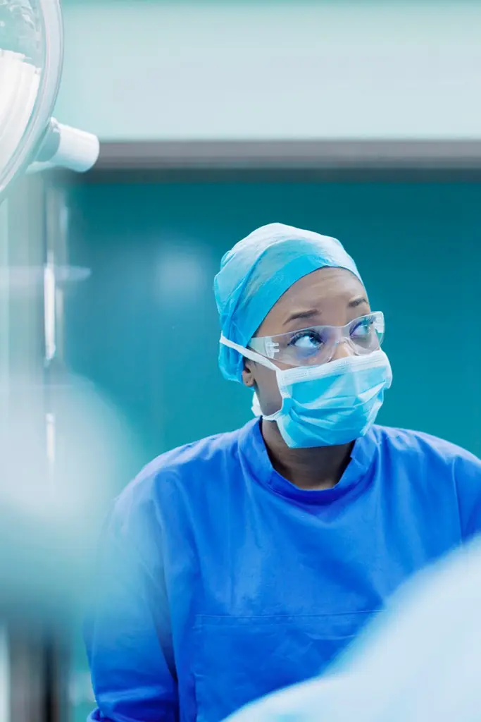 Bild einer Ärztin in blauer Kleidung in einem hellen Raum, mit durchsichtiger Brille, Haarschutz und Mund-Nasen-Maske.
