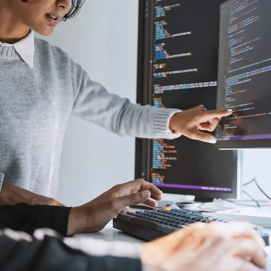 Referenzprojekt Corsience Corporate Design Relaunch Imagebild mit einer Frau in grauem Pullover, die auf einen von zwei Bildschirmen mit Computercode zeigt. Daneben sitzt eine Person in schwarzem Pullover, der etwas gezeigt wird.