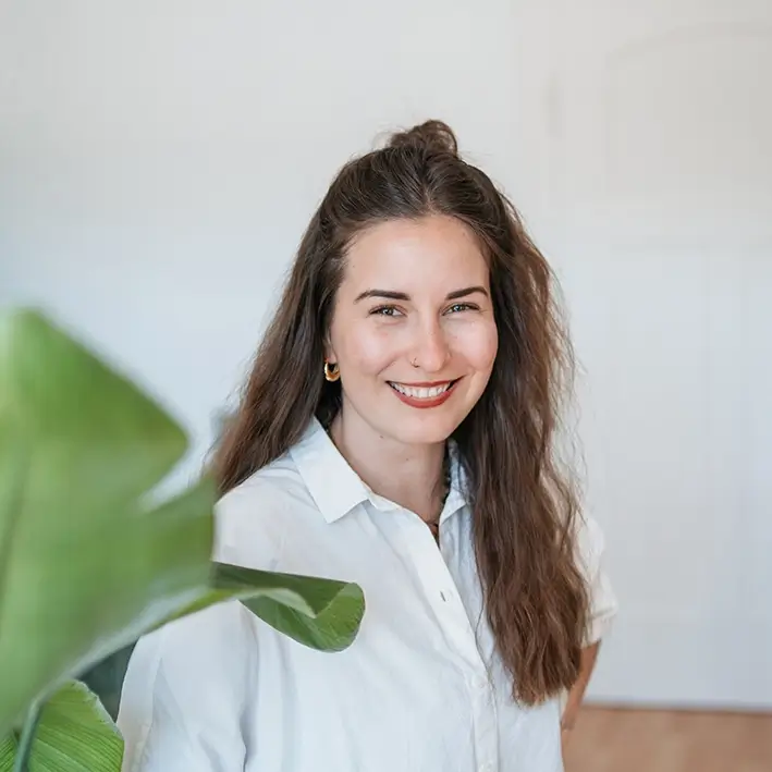 Porträt von junger Mitarbeiterin mit langen braunen Haaren im Vordergrund eine Pflanze Medical Communication bei Remy & Remy Healthcare Kommunikation