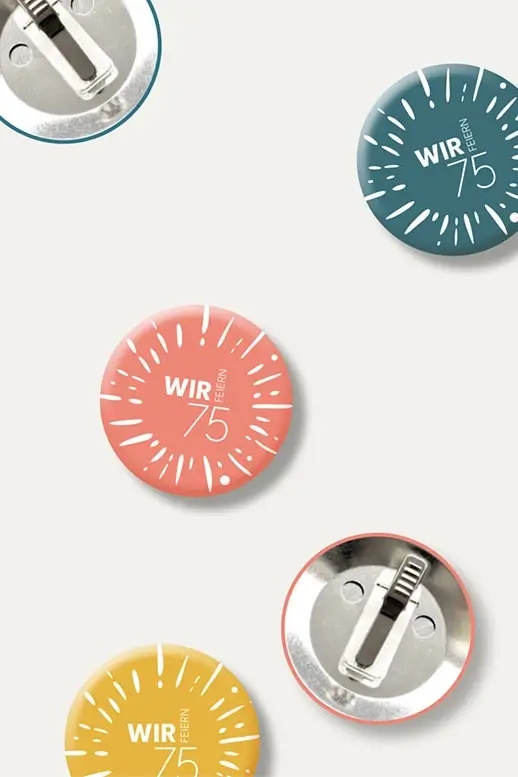 Referenzprojekt Caritas-Krankenhaus St. Josef Jubiläumskampagne - Mockups - Ansteckbuttons in Blau, Rosa und Gelb auf weißem Hintergrund mit weiß aufgedrucktem Kampagnenlogo. Zwei Buttons liegen umgedreht.
