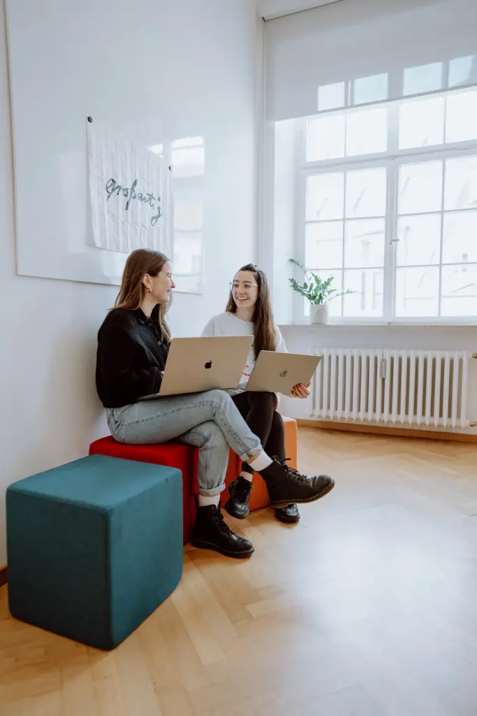 Zuständige für Medical Communication und Mitarbeiterin von Remy&Remy lachen während der Zusammenarbeit an einem Laptop im Agenturbüro und entwickeln im Gemeinschaftsraum auf bunten Sitzmöglichkeiten Konzepte für Healthcare-Marketing, Medical Content und Markenkommunikation.