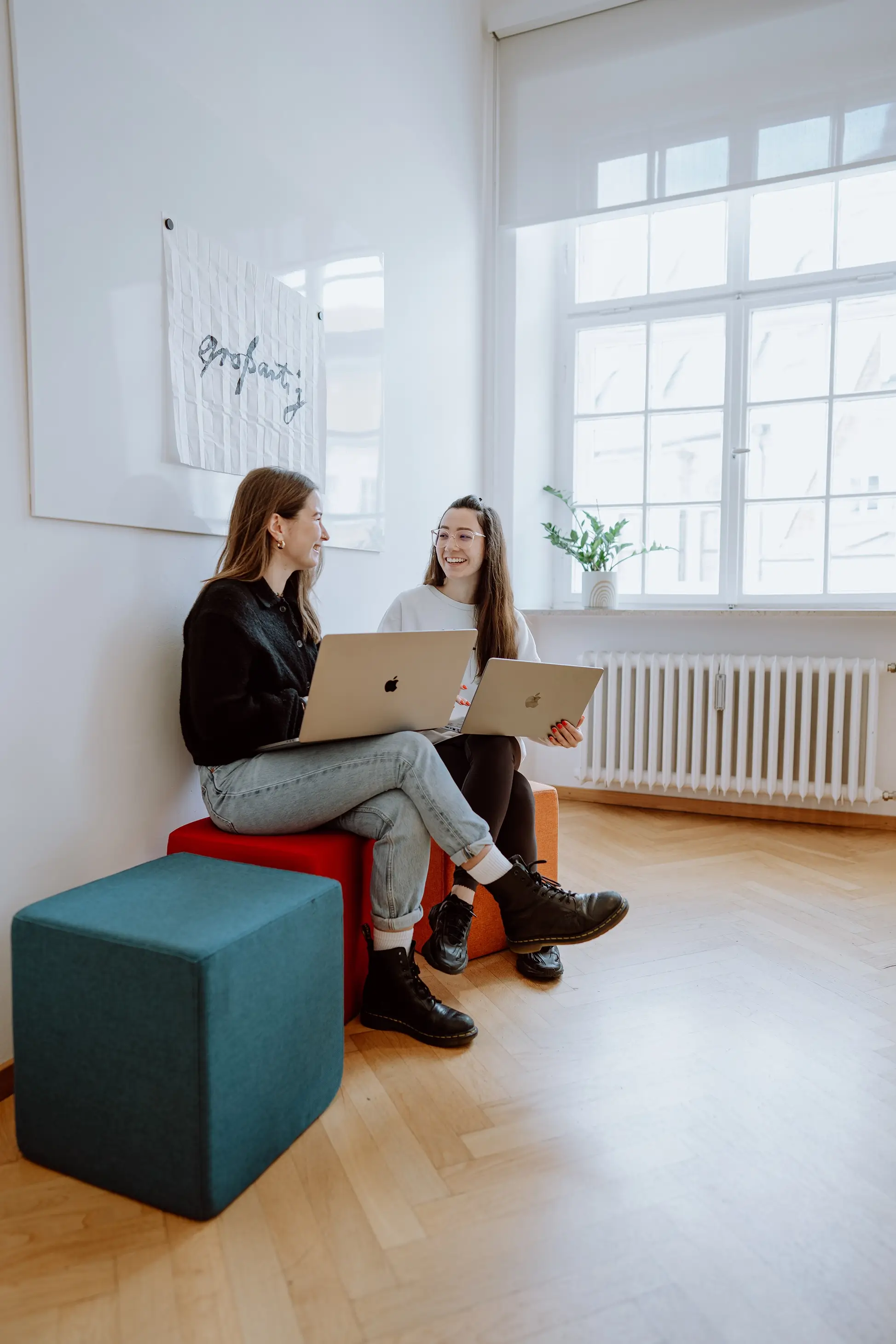 Zuständige für Medical Communication und Mitarbeiterin von Remy & Remy lachen während der Zusammenarbeit an einem Laptop im Agenturbüro und entwickeln im Gemeinschaftsraum auf bunten Sitzmöglichkeiten Konzepte für Healthcare-Marketing, Medical Content und Markenkommunikation.