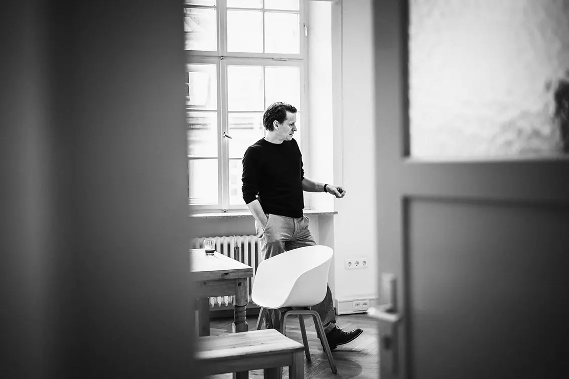 Geschäftsleitung von Remy & Remy mit dunklem Langarmshirt und Jeans steht im Gemeinschaftsraum, wird durch die Tür fotografiert und das Bild ist in Schwarz-Weiß dargestellt.