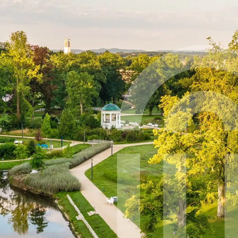 Referenzprojekt Bad Salzuflen - Bild vom Kurpark Bad Salzuflen, mit bunter Natur, großen Bäumen, gepflegten Rasenflächen, Wasserstelle, kleinem Häuschen und Sitzmöglichkeiten. Links im Bild, milchig-transparent, das Wasserzeichen.