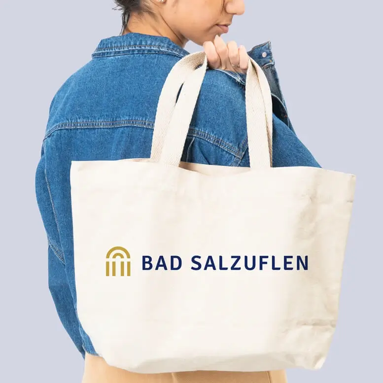 Referenzprojekt Bad Salzuflen - Mockup - Beige Einkaufstasche auf der das Logo abgebildet ist. Die Tasche wird von einer brünetten Frau gehalten, die eine Jeansjacke trägt.