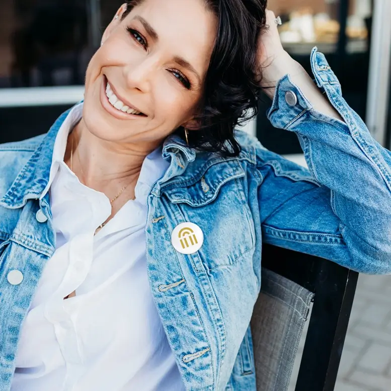 Referenzprojekt Bad Salzuflen Giveway Mockup in Form eines Ansteckbuttons auf dem das Logo abgebildet ist. Dieser wird von einer brünetten Frau getragen, die eine weiße Bluse und eine Jeansjacke trägt, an welcher der Ansteckbutton befestigt ist. Sie Lächelt glücklich.
