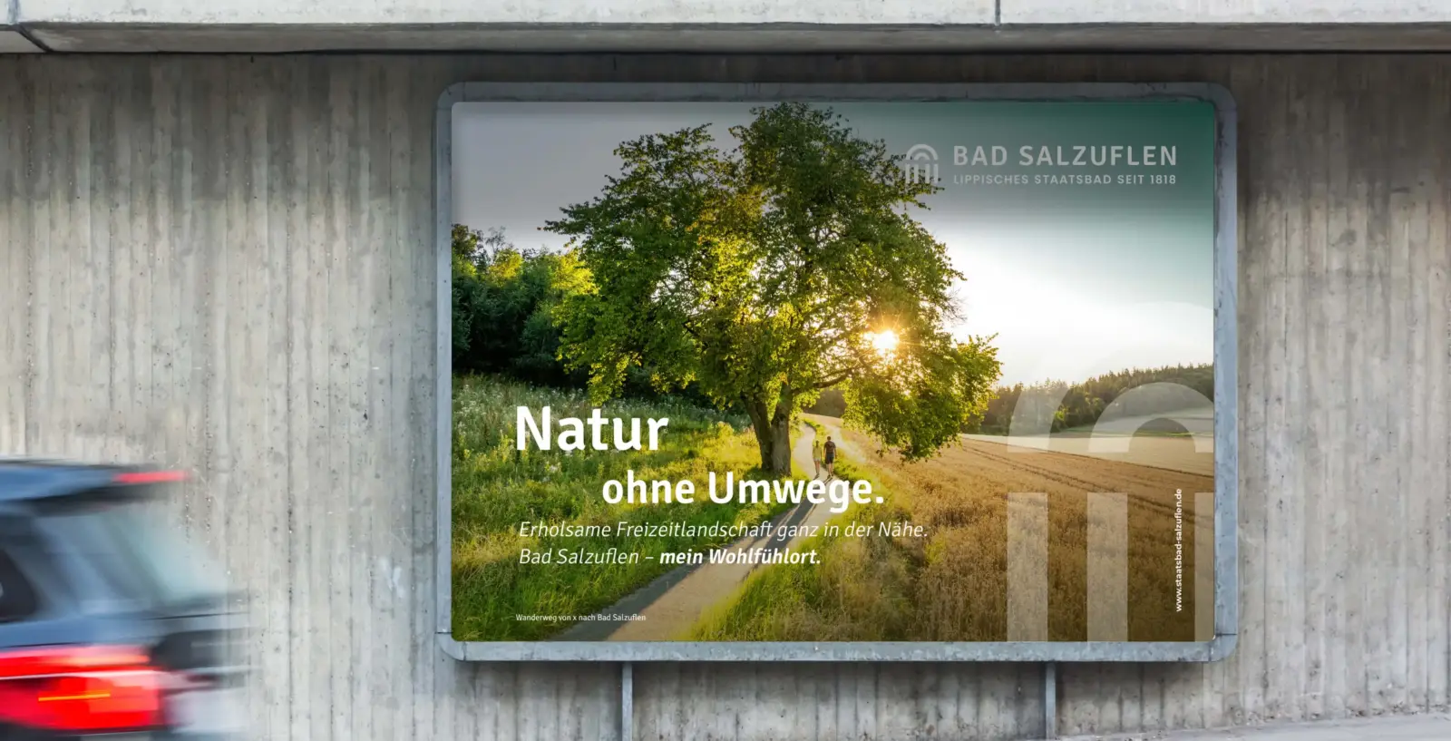 Referenzprojekt Bad Salzuflen Giveway Kampagne - Mockup eines Großflächenplakats an einer Straßenwand. Das Plakat zeigt einen großen Baum zwischen Feldern vor einem Sonnenuntergang und enthält einen Werbeslogan. Im Vordergrund fährt ein Auto vorbei, dessen Heck unscharf und verschwommen zu sehen ist.