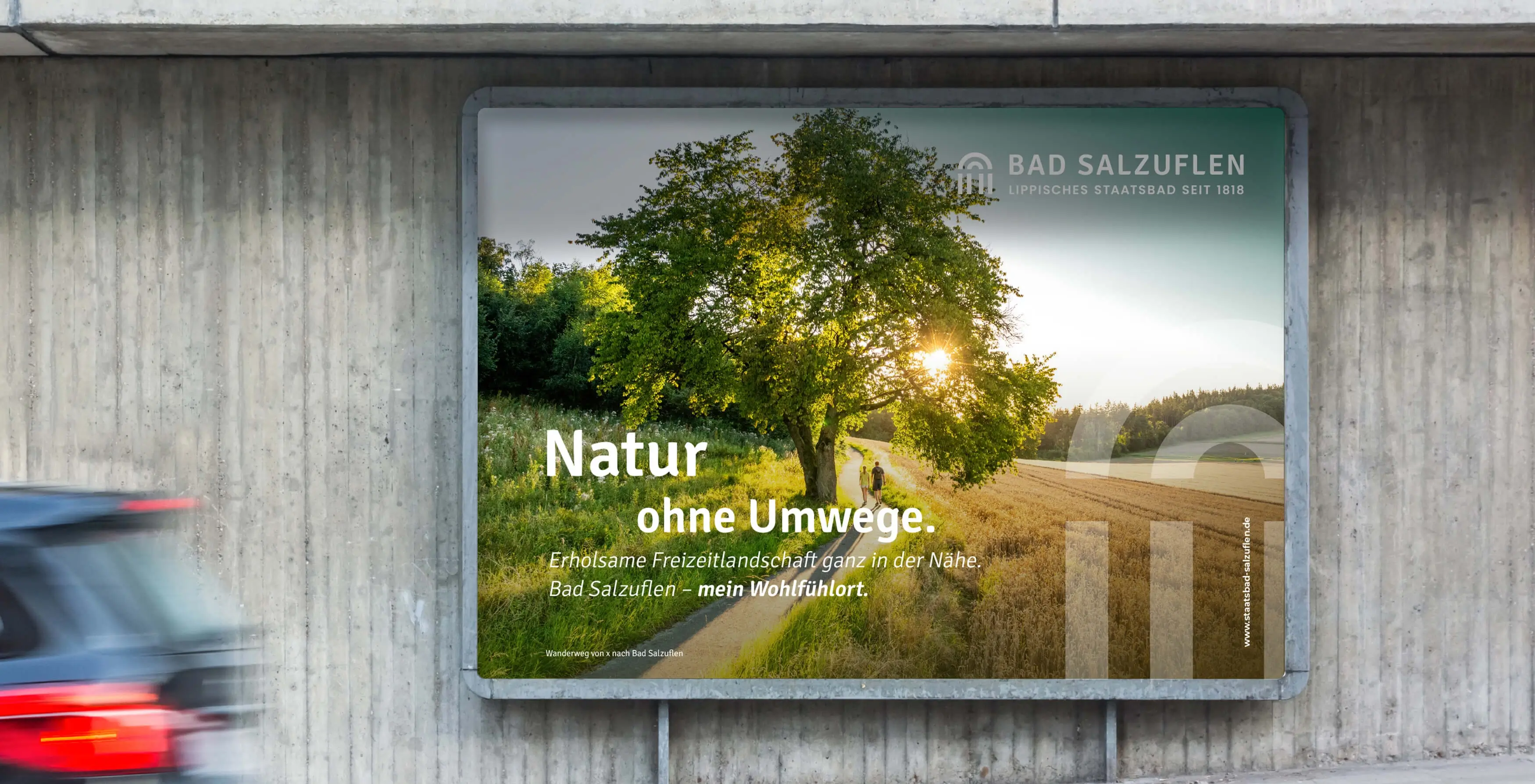 Referenzprojekt Bad Salzuflen Giveway Kampagne - Mockup eines Großflächenplakats an einer Straßenwand. Das Plakat zeigt einen großen Baum zwischen Feldern vor einem Sonnenuntergang und enthält einen Werbeslogan. Im Vordergrund fährt ein Auto vorbei, dessen Heck unscharf und verschwommen zu sehen ist.