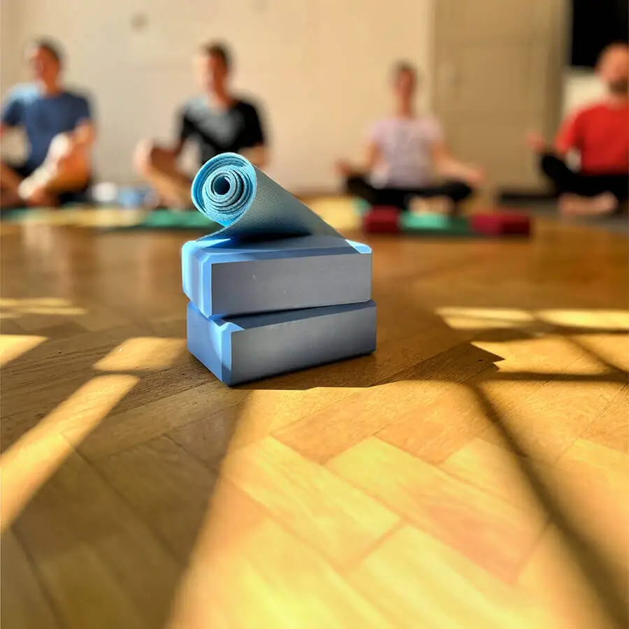 Yogamatte und Yogablöcke im Vordergrund, dahinter vier Personen beim Yoga im Gemeinschaftsraum der Agentur als Teil von betrieblichem Gesundheitsmanagement, Mitarbeitergesundheit und Workplace Wellbeing.