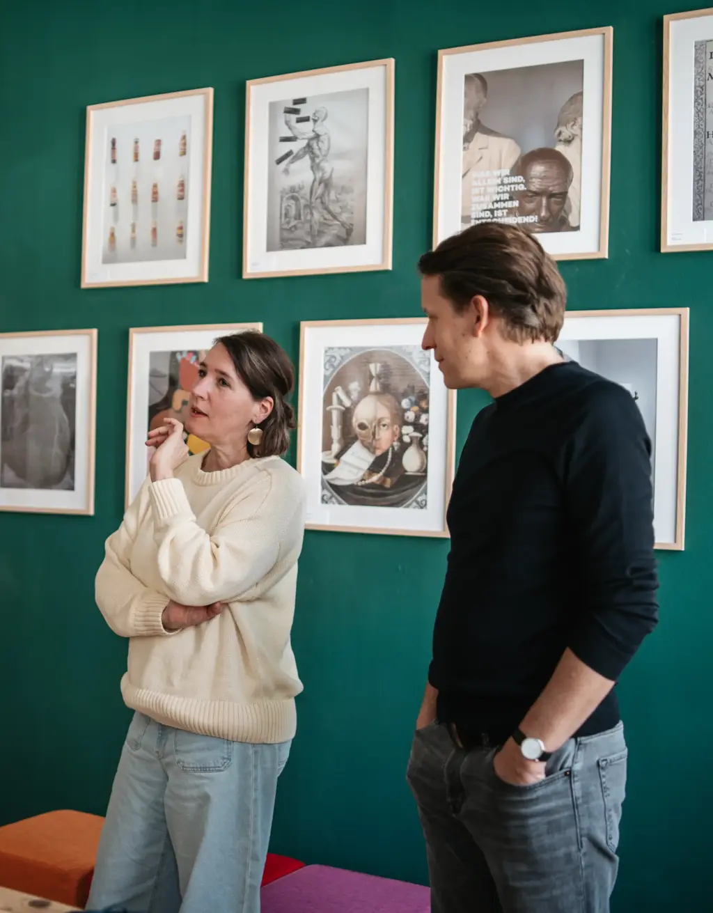 Eine Designerin der Agentur Remy&Remy im Gespräch im Gemeinschaftsraum mit bunten Sitzmöbeln und dekorierter Wandgestaltung, neben ihr steht einer der Geschäftsführer der Healthcare-Kommunikationsagentur.
